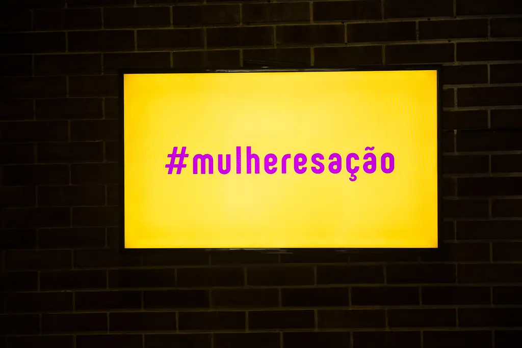 Painel iluminado exibe a hashtag #mulheresação em parede de tijolos