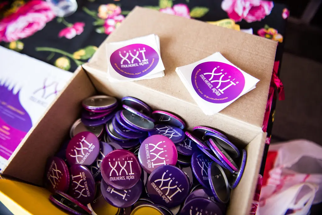 Adesivos e bótons roxos do festival organizados em caixa sobre mesa florida